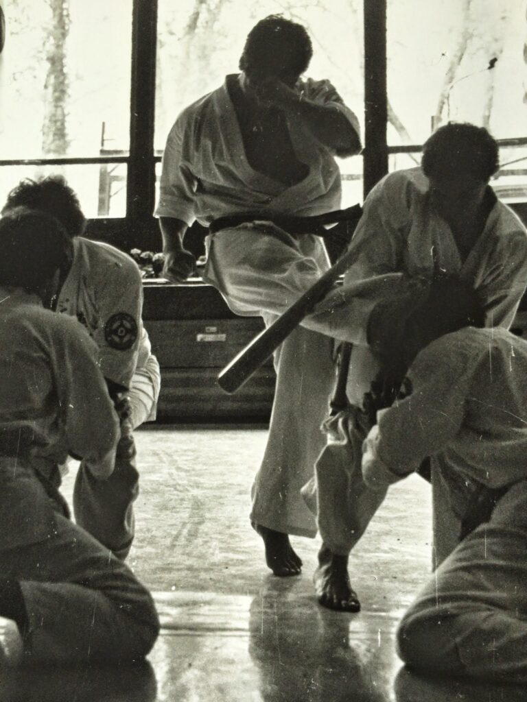 Dr. Bomi Joseph breaking a baseball bat with his shin on stage, supported by four karatekas in martial arts uniforms