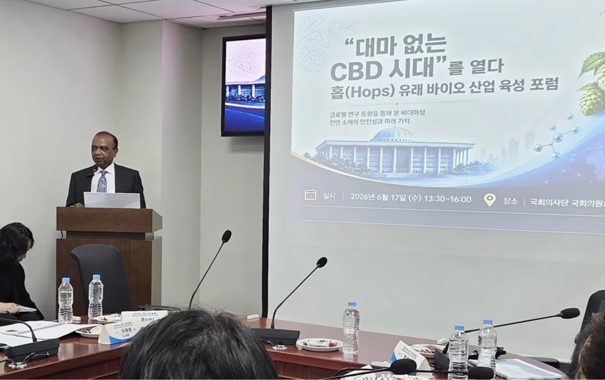 Dr. Bomi Joseph speaking at a podium during the Hops-Derived Bio-Industry Forum at the South Korean National Assembly Members' Hall, Seoul, with National Assembly Member Moon Jin-seok and other attendees visible in the audience.