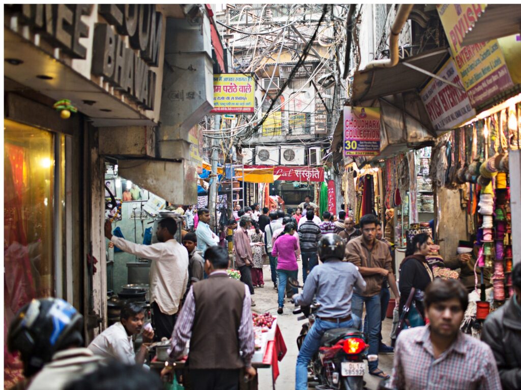 Busy side street shopping area in Delhi with pedestrians, small shops, vehicles, and uneven street conditions.